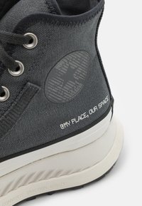Donkergrijze high-top sneaker met een gestructureerde canvas bovenkant, zwarte accenten, een cirkelvormig logo en witte tekst "@MY PLACE, OUR SPACE" op de zijkant.