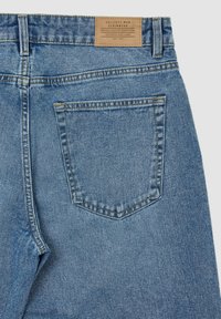 Jean en denim bleu clair avec une poche arrière unique, un léger délavage et une étiquette en cuir à la taille avec les détails de la marque.