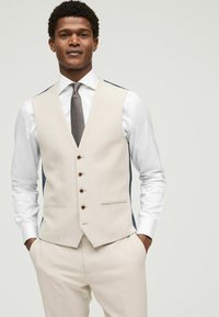 Gilet beige avec des panneaux latéraux marine, cinq boutons marron et deux poches avant, associé à une chemise blanche et une cravate à motifs. Coupe ajustée.
