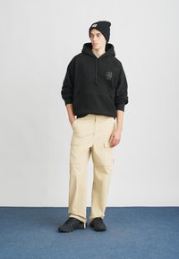 Μαύρο φούτερ με κουκούλα, μπροστινή τσέπη και λογότυπο, σε συνδυασμό με μπεζ παντελόνι cargo με πολλές τσέπες και μαύρα αθλητικά παπούτσια.
