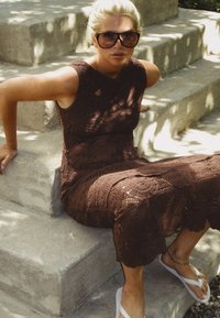 Abito all'uncinetto marrone con design senza maniche, con elaborati motivi floreali e orlo al di sotto del ginocchio. Infradito bianche e occhiali.