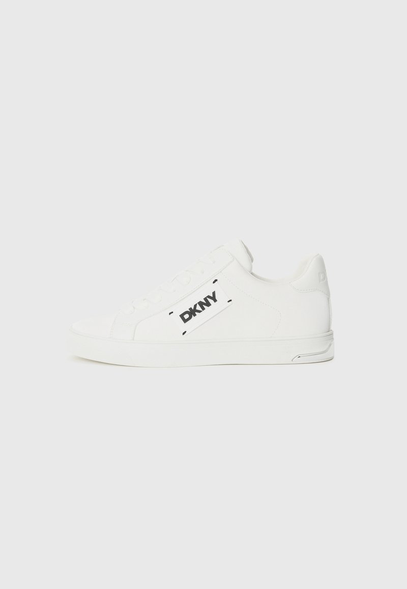 Bijele kožne teniske s ravnim potplatom, okruglim nosom i crnim DKNY logotipom sa strane. Glatka tekstura s minimalnim detaljima.