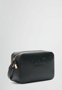 Sac à bandoulière noir en synthétique texturé avec une fermeture éclair et des accessoires en or, arborant un logo en relief sur le devant. Forme rectangulaire.