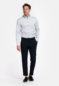 Chemise blanche à boutons avec un motif bleu discret, associée à un pantalon noir slim fit. Le modèle porte des chaussures noires à enfiler.