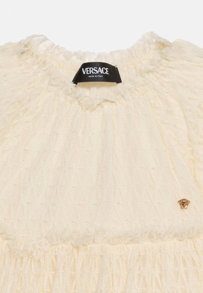 Εκρού διάφανο δαντελένιο ένδυμα με πιεσμένο λαιμόκοψη, μαύρη ετικέτα Versace και μικρό χρυσό έμβλημα Medusa στο κάτω δεξιά μέρος.