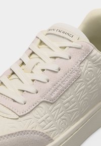 Κοντινό πλάνο αθλητικού παπουτσιού σε κρεμ χρώμα με ανάγλυφο μοτίβο λογότυπου Armani Exchange, πάνελ από σουέτ και λευκά κορδόνια περασμένα από τρύπες.