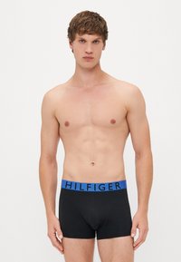 Ein oberkörperfreier Mann, der frontal steht und schwarze Boxershorts mit einem blauen Bund trägt, auf dem "HILFIGER" steht, vor einem weißen Hintergrund.