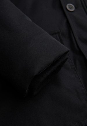 Close-up van een zwarte jas met een gladde stofstructuur, knoopdetail en opgerolde mouwen die een zachte voering laten zien.