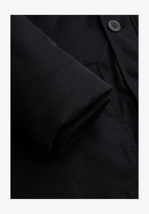 Close-up van een zwarte jas met een gladde stofstructuur, knoopdetail en opgerolde mouwen die een zachte voering laten zien.