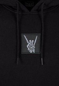 Crna hoodie s pravokutnom aplikacijom sivo-bijele kosturne ruke koja pravi znak. Mekana textura, vezice i opušteni kroj.