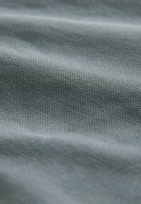 Tissu texturé dans un bleu-vert atténué, présentant un motif finement tissé et un drapé subtil, soulignant la douceur de la texture et la qualité du matériau.