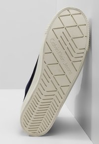 Valkoisen sneakerin pohja, jossa on geometrinen kuviointi ja "Calvin Klein Jeans" -brändäys, asetettu pystysuoraan valkoista kulmaa vasten.