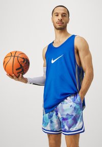 Sinine võrkpallide särk valge Nike logoga, koos lillemustriga lühikeste pükstega helesinisel ja violetset päritoluga. Käes oranž korvpall.