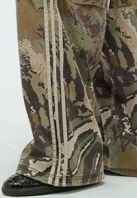 Camouflage wide-leg pants in shades of green, brown, and beige with three vertical beige stripes. Black textured shoes complete the look.