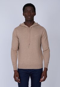 Hoodie marron clair en tissu doux, avec un cordon de serrage, des poignets côtelés et une coupe ajustée. Porté sur un jean bleu foncé.