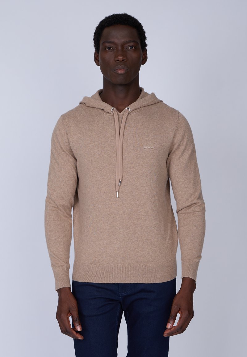 Hoodie marron clair en tissu doux, avec un cordon de serrage, des poignets côtelés et une coupe ajustée. Porté sur un jean bleu foncé.