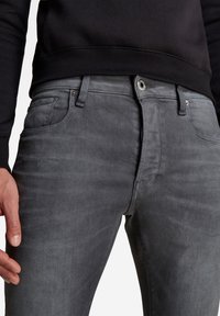 Šedé džínové kalhoty s úzkým střihem, které mají klasický design s pěti kapsami, stříbrný knoflík a jemné vyblednutí po celé látce.