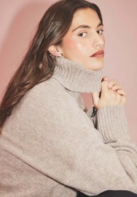 Helles beige Rollkragenpullover mit geripptem Muster und weiten Ärmeln, die mit gefalteten Händen getragen werden. Glattes, weiches Material vor einem rosafarbenen Hintergrund.