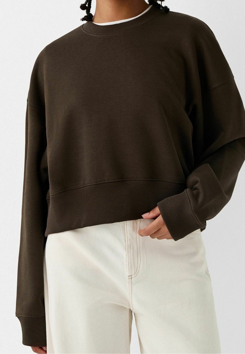 Brun oversized sweatshirt med ribbad nederkant och ärmar, med rund halsringning och mjukt, slätt material. Kombinerad med ljusa byxor.