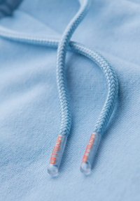 Hoodie bleu clair avec des cordons tressés dotés d'extrémités en plastique transparent, mis en valeur par du texte orange. Texture de tissu lisse et douce visible.