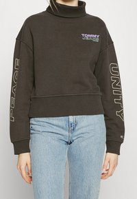 Persona che indossa un felpa nera a collo alto con la scritta "Tommy Jeans", "Pace" e "Unità", abbinata a jeans in denim azzurro chiaro.