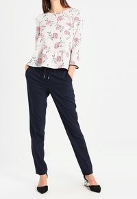 Blusa crema con fantasia floreale e maniche a tre quarti, abbinata a pantaloni slim-fit blu navy e scarpe nere con punta affusolata.