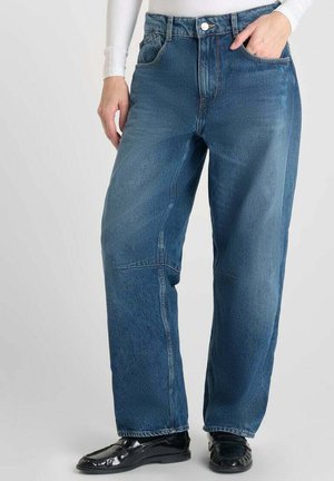 Personne portant un jean en denim bleu ample avec des poches avant, un bouton et une fermeture éclair, associé à des mocassins noirs et un haut à manches longues blanc.