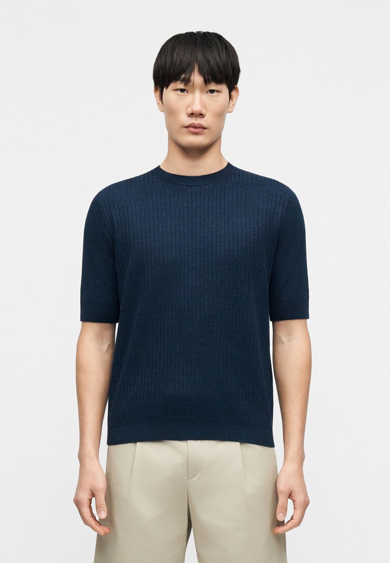 Pull en maille côtelée bleu marine à manches courtes avec un col rond, un tissu texturé et un ourlet droit ; porté avec un pantalon de couleur claire.