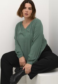 Blouse verte texturée avec un col en V et des accents de boutons, associée à un pantalon noir taille haute et des bottines. Design simple et décontracté.