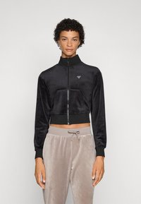 Juoda trumpa zip-up striukė, pagaminta iš minkštos medžiagos, su aukštu apykakle, turinti priekinį kišenę ir mažą logotipą ant krūtinės.