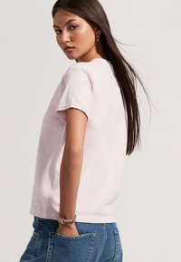 Svijetloružičasta pamučna majica kratkih rukava i opuštenog kroja, u kombinaciji s plavim trapericama. Na rukavima su vidljivi detalji manžeta.