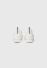 White athletic shoes with textured mesh upper, rubber sole, and a gold accent on the pull tab. Features a raised logo on the heel.