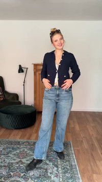 Νέα γυναίκα μέσα σε εσωτερικό χώρο, φοράει μια ναυτική ζακέτα, λευκή μπλούζα, μπλε τζιν, μαύρα μποτάκια και γυαλιά ηλίου στο κεφάλι, και στέκεται σε ένα σχέδιο χαλί.