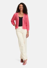 Cardigan en crochet corail à mailles ouvertes, à manches trois-quarts et coupe décontractée, associé à un pantalon large crème et des talons roses.