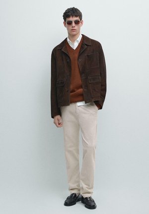 Brun ruskindsjakke, rustfarvet sweater, hvid skjorte, beige bukser og sorte loafers. Model iført mørke solbriller. Glatte teksturer og lagdelt design.