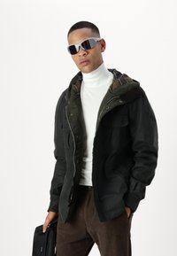 Black jacket with a green, plaid-lined hood, worn over a white turtleneck. The model wears gray sunglasses and holds a black bag.
