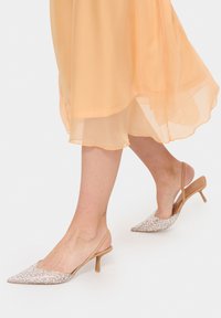 Beige slingback-klackar med spetsig tå, med ett glittrande silveröverdrag och en tunn, elegant klack. Bärs med en fladdrande, genomskinlig persikofärgad klänning.