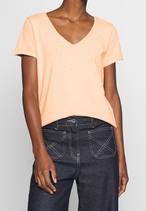 Vrouw draagt een licht perzikkleurige V-hals top met korte mouwen, ingestopt in een donkerblauwe jeans met hoge taille en witte stiknaden op de voorzakken.