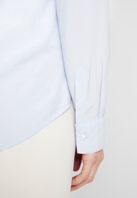 Camicia a maniche lunghe azzurro chiaro con una texture liscia, caratterizzata da un polsino puntato con bottone e un orlo sagomato.