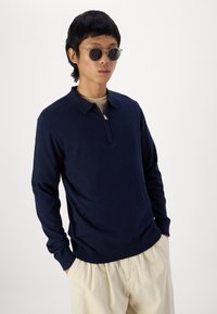 Pullover navy a maniche lunghe con colletto zip e polsini a costine. Indossato sopra una camicia chiara, abbinato a pantaloni chiari e occhiali da sole.