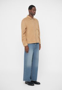 Tan wool blend jacket with a collar and front pocket, paired with wide-leg blue denim jeans that fade to lighter shades. Black shoes.