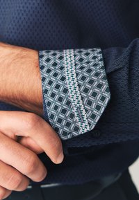 Blå skjorteknap med mønstret design i geometriske figurer i nuancer af blågrøn og hvid, med en kontrasterende kant langs kanten.