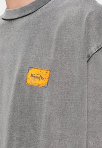 Γκρι βαμβακερό μπλουζάκι με στρογγυλή λαιμόκοψη και κατεβασμένους ώμους, με ένα φθαρμένο κίτρινο ταμπελάκι με την ένδειξη "Replay Inc." στο στήθος.