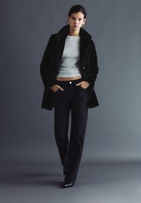 Femeie purtând o haină neagră din blană artificială peste un top gri scurt, blugi întunecați cu model, cizme negre ascuțite, mergând cu mâinile în buzunare.