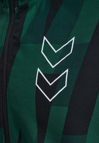 Groene en zwarte zip-up jack met een geometrisch patroon, gestructureerde stof en twee witte chevron-logo's op de borst.