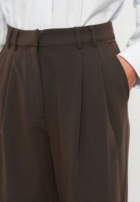 Pantaloni marroni su misura con pieghe, una texture liscia e tasche laterali. Vita aderente con passanti per cintura, abbinati a una camicia bianca.