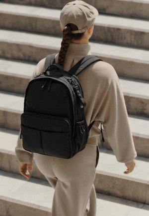 Person iført beige tøj og kasket, bærende på en sort rygsæk, går op ad udendørs stentrapper.