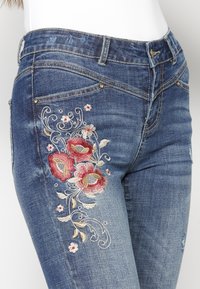 Denimjeans med intrikata blommönster i röd och krämfärg på vänster lår, med en medeltvätt och lätt blekning.