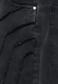 Μαύρο τζιν ύφασμα με τέσσερις παράλληλες ξεσκισμένες ραφές και ασημένιο μεταλλικό κουμπί κοντά στην άκρη της τσέπης.