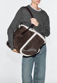Person wearing grey sweater and jeans holding large brown shearling tote bag with dark handles over shoulder.